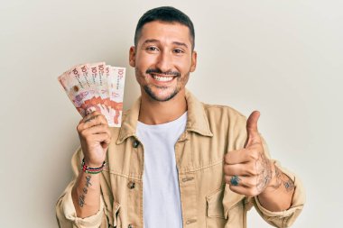 Elinde 10 tane Kolombiya pezosu olan yakışıklı adam mutlu ve olumlu gülümsüyor, başparmağıyla mükemmel ve onay işareti yapıyor. 