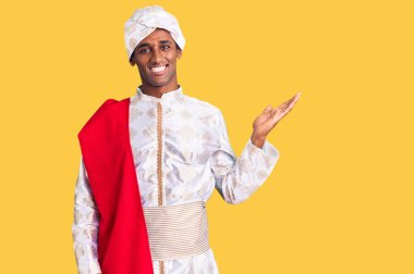 Gelenek giyinmiş Afrikalı yakışıklı bir adam Sherwani sari giysileri giymiş neşeli bir şekilde gülümseyen ve avuç içiyle kameraya bakan.. 