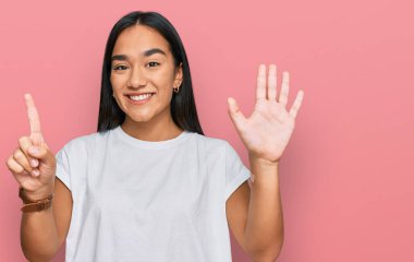 Genç Asyalı kadın, günlük beyaz tişört giyip altı numaralı parmaklarıyla işaret ederken kendinden emin ve mutlu gülümsüyor.. 