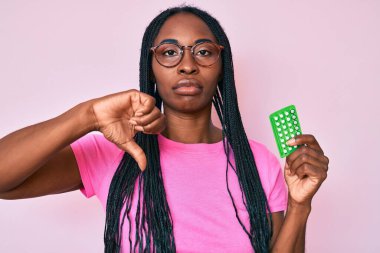 Örgülü Afrikalı Amerikalı kadın elinde kızgın yüzlü doğum kontrol hapları, olumsuz işaretler baş parmakları aşağıda, reddedilme konsepti var. 