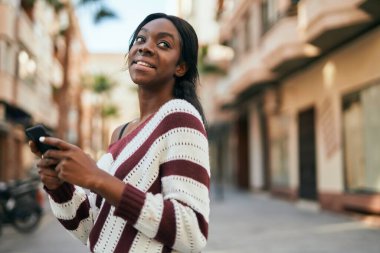 Genç Afrikalı Amerikalı kadın şehirde akıllı telefon kullanarak mutlu bir şekilde gülümsüyor..