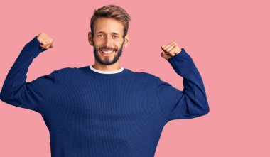 Sakallı, sıradan süveter giyen yakışıklı sarışın adam gururla gülümseyen kol kaslarını gösteriyor. fitness konsepti. 