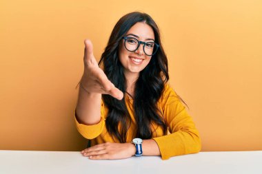 Güzel, esmer, gözlüklü ve gündelik kıyafetleri olan genç bir kadın masada oturmuş dostça gülümseyerek selamlaşıp selamlaşıyorlar. başarılı iş. 