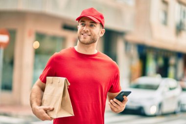 Beyaz atlı genç teslimatçı akıllı telefon kullanıyor ve şehirde kağıt torba taşıyor..