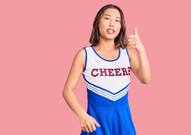 Genç, güzel Çinli kız ponpon kız üniforması giyip mutlu bir şekilde el hareketi yapıyor. Kameraya bakıp başarıyı gösteren ifade. 