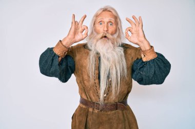 Gri saçlı, uzun sakallı, Viking kostümü giyen yaşlı bir adam. Şaşırmış ve şaşırmış görünüyor. Tamam sembolünü parmaklarıyla yapıyor. çılgın ifade 