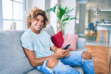 Genç İspanyol adam evde akıllı telefon kullanarak mutlu bir şekilde gülümsüyor.