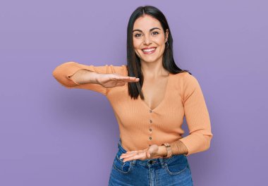 Latin kökenli genç bir kadın, gündelik kıyafetler giyiyor. Elleriyle işaret ediyor. Ölçü sembolü. Kameraya bakıp gülümsüyor. ölçüm kavramı. 