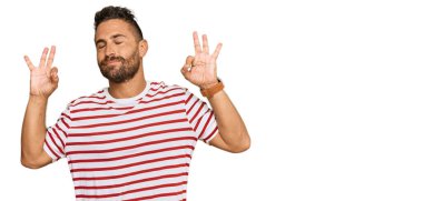 Sakallı, çizgili tişörtlü yakışıklı adam rahatla ve gözleri kapalı gülümse parmaklarıyla meditasyon hareketi yapıyor. yoga konsepti. 