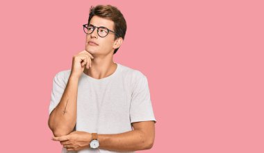 Sıradan kıyafetler giyen yakışıklı beyaz bir adam ve eli çenesinde gözlük takan soru soran, düşünceli bir ifade. Gülümseyen ve düşünceli bir yüz. şüphe kavramı. 