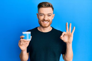 Kulak kiri temizleyici tutan genç kızıl saçlı adam parmaklarıyla imza atıyor, gülümseyerek mükemmel bir sembol oluşturuyor. 