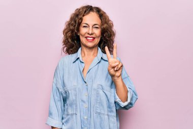 Orta yaşlı güzel bir kadın pembe arka plan üzerinde sıradan bir kot gömlek giyiyor gülümsüyor parmaklarını zafer işareti yaparken gösteren kameraya bakıyor. İki numara..