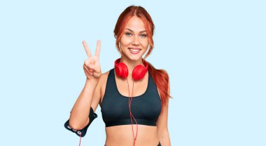 Spor kıyafetleri giyen, kulaklık takan, kameraya gülümseyen ve zafer işareti yapan kızıl saçlı genç bir kadın. İki numara.. 