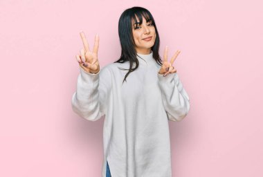 Kâküllü, balıkçı yaka kazak giyen genç esmer kadın kameraya bakıp zafer işareti yaparken gülümsüyor. İki numara.. 