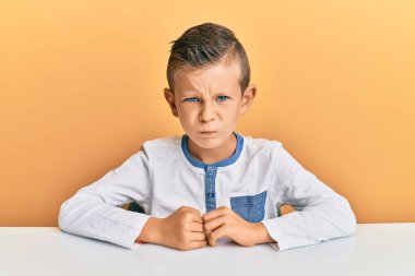 Kafkasyalı sevimli bir çocuk, gündelik giysiler giymiş şüpheci ve gergin bir şekilde masada oturuyor, sorun yüzünden suratını asıyor. negatif kişi. 