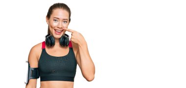 Güzel sarışın kadın spor kıyafetleri giyiyor ve kulaklık takıyor. Yüzüne ve burnuna parmak işareti yapıyor. Neşeli gülümsüyor. güzellik konsepti 