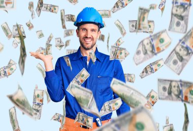 Genç, yakışıklı, işçi üniforması giyen ve kasketli neşeli bir şekilde gülümseyen, avuç içi kameraya bakan bir adam..