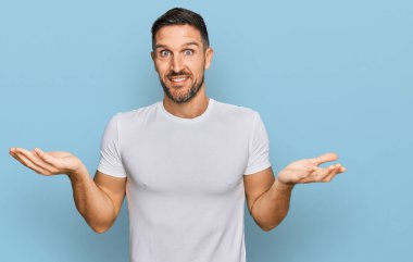 Sakallı, sıradan beyaz tişörtlü, kafası karışık ve kolları ve elleri havada yakışıklı bir adam. şüphe kavramı. 