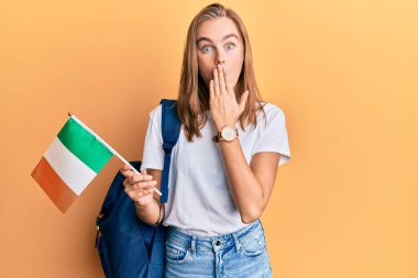 Güzel sarışın değişim öğrencisi elinde İrlanda bayrağı ile ağzı kapalı, şok olmuş ve hata yapmaktan korkan bir öğrenci. şaşırmış ifade 