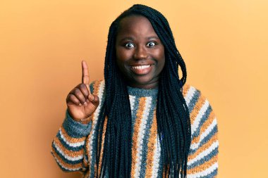 Saçları örgülü genç siyahi bir kadın sıradan bir kazak giyiyor ve bir numaralı parmağını gösterip gülümserken kendinden emin ve mutlu.. 