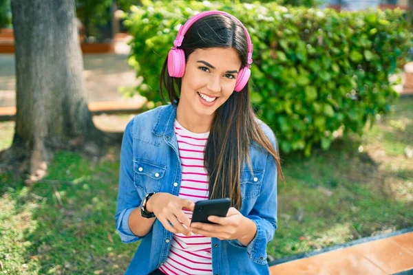 公園に座っているスマートフォンやヘッドフォンを使用して幸せな笑顔若い美しいヒスパニック系の女の子 — ストック写真