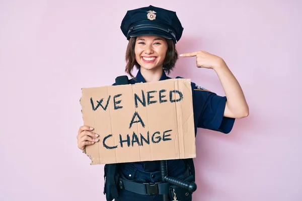Young beautiful woman wearing police uniform holding we need a change banner pointing finger to one self smiling happy and proud