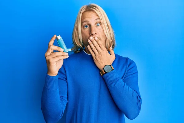 Caucasian young man with long hair holding medical asthma inhaler covering mouth with hand, shocked and afraid for mistake. surprised expression