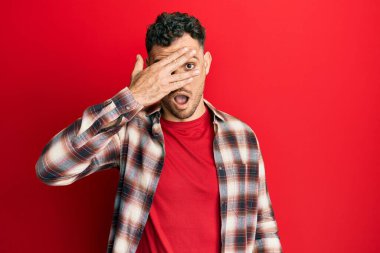 Gündelik kıyafetler giyen genç İspanyol adam şok içinde yüzünü ve gözlerini eliyle kapatıyor, parmaklarından korkuyla bakıyor. 
