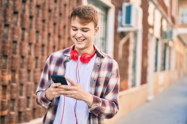 Genç beyaz adam şehirde akıllı telefon ve kulaklık kullanarak mutlu bir şekilde gülümsüyor..