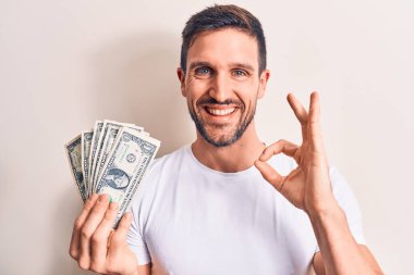 İzole edilmiş beyaz arka planda bir deste para tutan yakışıklı genç adam parmaklarıyla imza atıyor, gülümsüyor ve mükemmel bir sembol oluşturuyor.