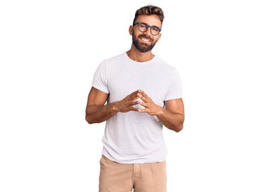 Gündelik kıyafetler ve gözlük takan genç İspanyol adam ellerini birleştirip gülümseyerek gevşemiş ve neşeliydi. Başarı ve iyimserlik 