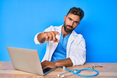 Klinikte çalışan, doktor üniforması giyen, kamerayı işaret eden genç bir İspanyol. Kendine güvenen, ciddi görünen bir jest. 