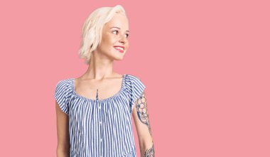 Dövmeli genç sarışın kadın. Sıradan kıyafetler giyiyor. Yüzünde gülümsemeyle yan yana bakıyor. Kendinden emin gülen. 