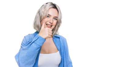 Sıradan kıyafetler giyen genç sarışın kız elinle mutlu bir jest yapıyor. Kameraya bakıp başarıyı gösteren ifade. 