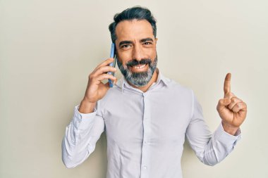 Orta yaşlı sakallı ve gri saçlı bir adam akıllı telefondan konuşuyor. Bir fikir veya soruyla gülümsüyor ve mutlu bir yüzle işaret ediyor. 