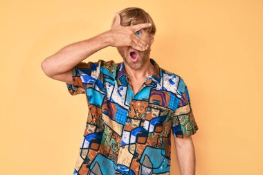 Young blond man wearing summer shirt peeking in shock covering face and eyes with hand, looking through fingers with embarrassed expression. 