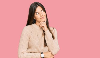 Genç esmer kadın, sıradan bir kazak giyiyor, elleri çenesinde soru soruyor, düşünceli bir yüz ifadesi var. Gülümseyen ve düşünceli bir yüz. şüphe kavramı. 