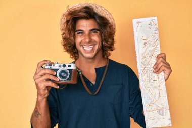 Klasik fotoğraf makinesi ve şehir haritası tutan genç İspanyol adam mutlu ve havalı bir gülümsemeyle gülümsüyor. diş gösterme. 