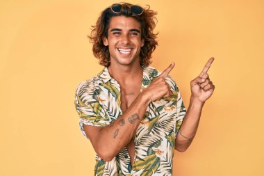 Yazlık tişört giyen genç İspanyol adam gülümseyip kameraya bakıyor ve iki eliyle ve parmaklarıyla kenarı işaret ediyor.. 