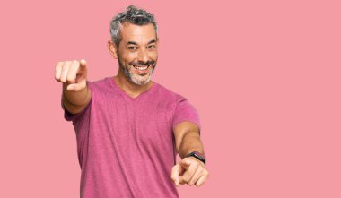 Orta yaşlı, gri saçlı, günlük kıyafetler giyen, seni ve kamerayı işaret eden, gülümseyen, pozitif ve neşeli bir adam. 