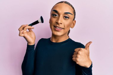 Makyaj yapan, elinde makyaj fırçası tutan, gülümseyen, mutlu ve olumlu gülümseyen, başparmağını kaldıran, mükemmel ve onay işareti yapan İspanyol bir adam. 