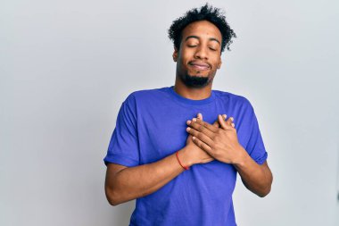 Sakallı, mavi tişörtlü, göğsünde elleri göğsünde, yüzünde minnet dolu bir jestle gülümseyen Afro-Amerikan bir genç. sağlık kavramı. 