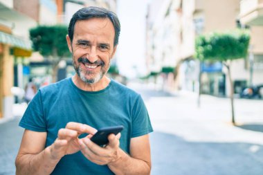 Sakallı orta yaşlı bir adam dışarıda mutlu bir şekilde gülümsüyor akıllı telefon kullanıyor.