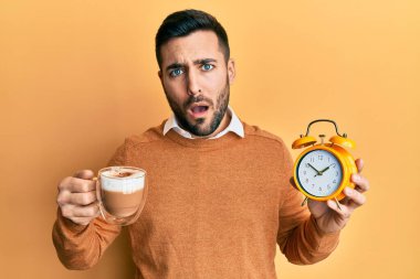 Young hispanic man holding coffee and alarm clock in shock face, looking skeptical and sarcastic, surprised with open mouth 