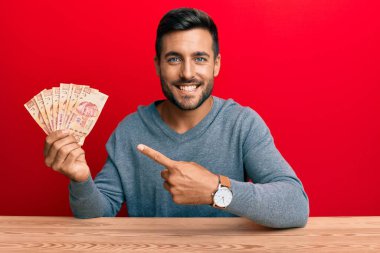 Meksika pezosu tutan yakışıklı İspanyol adam mutlu bir şekilde el ve parmak işareti yapıyor. 