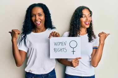 Afrika kökenli Amerikalı anne ve kız ellerinde kadın hakları pankartı başparmağıyla yan tarafa bakıyor mutlu bir şekilde gülümsüyor. 