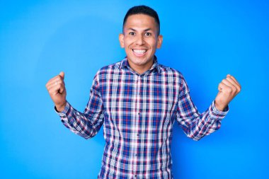 Gündelik giysiler giymiş genç, yakışıklı Latin bir adam kollarını kaldırıp, gözleri açık bir şekilde başarıyı kutluyor. Kazanan konsept. 