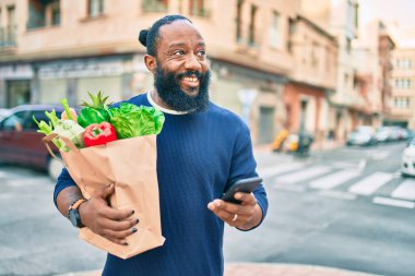 Sakallı Afrikalı Amerikalı adam elinde süpermarketten alınmış kağıt torbalarla akıllı telefon kullanıyor.