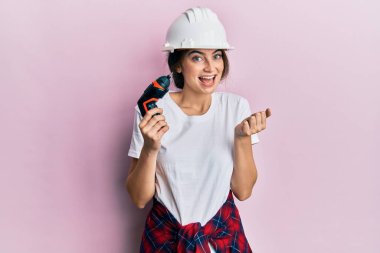 Genç beyaz kadın mimar şapkası takıyor. Matkap kullanıyor. Gururla bağırıyor, zaferi kutluyor ve başarıyı kutluyor. 