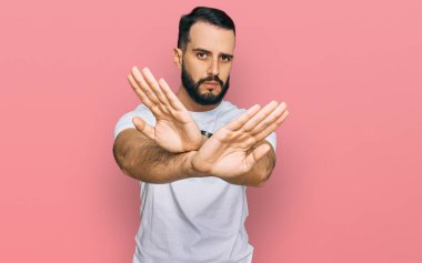 Sakallı genç adam, günlük beyaz tişört giyiyor. Reddedilmiş yüz ifadesi, kollar çapraz, avuç içleri negatif işareti, kızgın yüz ifadesi. 